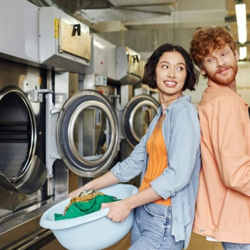 imgi_65_smiling-multiethnic-couple-standing-back-to-back-and-holding-basins-with-clothes-in-coin-laundry-e1716535733496